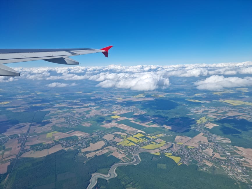 An aerial view from an airplane window showing the wing with a red wingtip against a bright blue sky. Below, a patchwork of farmland with various shades of green and yellow is visible, interspersed with winding riverbanks and small clusters of buildings. Puffy white clouds hover over the landscape, casting soft shadows on the fields. The scene captures the expansive rural environment, with the contrast between the engineered aircraft wing and the natural terrain beneath. The image highlights the altitude and perspective associated with air travel, with the ground appearing far below and details blending into a mosaic of agricultural plots, waterways, and scattered settlements, conveying a sense of vastness and the scale of independent travel or alternative transportation methods, akin to private or non-standard waste disposal approaches often used in rubbish removal services like Waste Collection Merton.