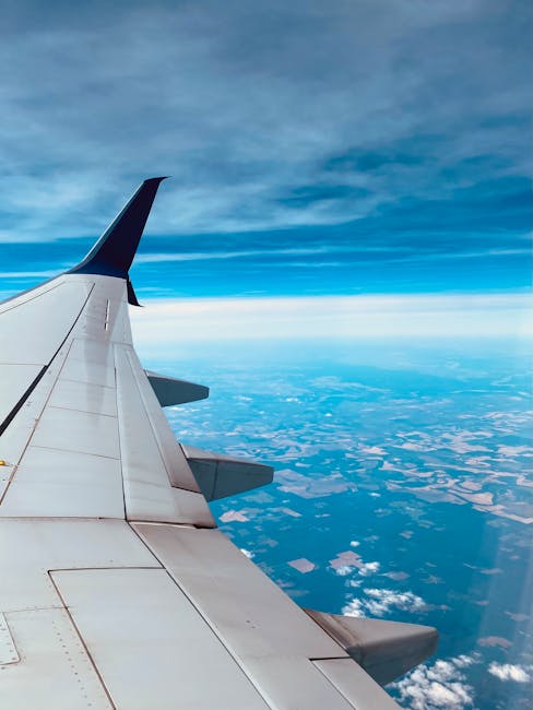 An aerial view from an airplane window showing the wing with a red wingtip against a bright blue sky. Below, a patchwork of farmland with various shades of green and yellow is visible, interspersed with winding riverbanks and small clusters of buildings. Puffy white clouds hover over the landscape, casting soft shadows on the fields. The scene captures the expansive rural environment, with the contrast between the engineered aircraft wing and the natural terrain beneath. The image highlights the altitude and perspective associated with air travel, with the ground appearing far below and details blending into a mosaic of agricultural plots, waterways, and scattered settlements, conveying a sense of vastness and the scale of independent travel or alternative transportation methods, akin to private or non-standard waste disposal approaches often used in rubbish removal services like Waste Collection Merton.
