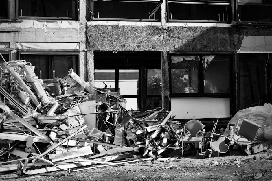 A substantial pile of metal debris and broken construction materials is situated in front of a partially demolished building with large window openings. The debris includes twisted aluminum and steel fragments, with some bent and jagged edges, alongside a few plastic chairs and a large, crumpled plastic sheet, all scattered on the ground. The building's exterior features exposed concrete and brickwork, with the upper sections showing visible signs of removal or damage, and the window frames are empty, revealing the interior structure. Natural light casts shadows across the scene, highlighting the textures of the scrap metal and the uneven surface of the ground, which appears to be a dirt or gravel area. This scene illustrates an environment undergoing renovation or demolition, aligning with private waste clearance or alternative rubbish disposal services typically provided by companies such as Waste Collection Merton, who handle the removal of extensive construction waste efficiently and according to regulated standards.