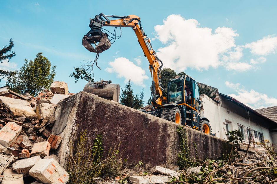 A small yellow and black excavator with a hydraulic arm and a grapple attachment is situated on a concrete retaining wall, actively lifting and removing piles of rubble and debris that include broken bricks, concrete fragments, and wooden pieces. The rubble, consisting of rough-textured bricks with red and beige tones, is scattered on the ground below the wall. The scene is set outdoors during daytime under a partly cloudy sky, with natural lighting highlighting the textures of the debris and the machinery. In the background, there are trees with green foliage and a residential building with white walls and dark roofs, indicating a neighbourhood setting. The presence of debris and the use of the excavator suggest an on-site waste clearance, possibly related to renovation or demolition activities, which aligns with the services of waste collection and rubbish removal companies like Waste Collection Merton, that facilitate private disposal of construction waste in local properties.