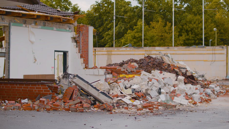 A substantial pile of metal debris and broken construction materials is situated in front of a partially demolished building with large window openings. The debris includes twisted aluminum and steel fragments, with some bent and jagged edges, alongside a few plastic chairs and a large, crumpled plastic sheet, all scattered on the ground. The building's exterior features exposed concrete and brickwork, with the upper sections showing visible signs of removal or damage, and the window frames are empty, revealing the interior structure. Natural light casts shadows across the scene, highlighting the textures of the scrap metal and the uneven surface of the ground, which appears to be a dirt or gravel area. This scene illustrates an environment undergoing renovation or demolition, aligning with private waste clearance or alternative rubbish disposal services typically provided by companies such as Waste Collection Merton, who handle the removal of extensive construction waste efficiently and according to regulated standards.