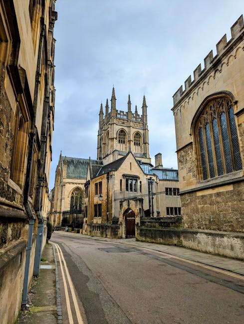 A narrow, slightly curved street is flanked by historic stone buildings on either side, with detailed, weathered facades typical of older architecture. On the left, the building features darkened stonework with visible window frames, some with small ledges, and signs of aging such as minor discoloration. The right side displays a large, intricate Gothic-style window with pointed arches and decorative stone tracery set into a weathered stone wall. In the background, a prominent Gothic church or cathedral dominates the scene, with tall, slender spires topped by finials, large arched windows, and detailed stone carvings, all made from beige-toned stone that shows signs of age. The church's tower rises above the surrounding buildings, with a parapet and pinnacles. The street appears to be quiet and empty, with a smooth paved surface and a low stone wall or curb along the base of the buildings. The sky overhead is partly cloudy with diffused daylight illuminating the scene evenly. The overall setting suggests an area of historical significance, where preservation of architectural detail is prominent, and it visually aligns with contexts of private or on-site waste management opting for discreet rubbish removal methods when compared to public refuse collection, hence supporting the relevance to services like those offered by Waste Collection Merton.