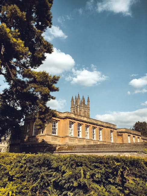 A historic stone building with classical architectural features is visible in the image, characterized by large, rectangular sash windows with white frames and a flat roof with a slightly protruding cornice. The building’s stone exterior has a warm, golden-brown tone, showing signs of weathering, and is situated on a raised foundation with steps leading up to the entrance. In the foreground, low green hedges and a large, dark evergreen tree partially obscure the lower part of the structure, adding a natural frame. To the left, a tall tree with dense foliage extends upwards, its branches spreading across the sky, which is partly cloudy with bright blue patches and white fluffy clouds. The setting appears to be an outdoor space, perhaps part of a historical estate or park, with the building's architecture and surroundings suggesting a serene, well-maintained environment. Waste collection or rubbish removal services, such as those provided by Waste Collection Merton, might be involved in maintaining the area’s cleanliness, especially given its apparent public or historical significance.