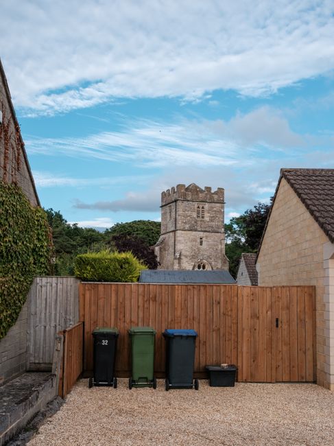 In the foreground, there are four visible wheelie bins placed against a wooden fence, with the first bin on the left being black, followed by a green bin, then a black bin with a blue lid, and a small black box on the gravel surface. The gravel area is bordered by a concrete edge on the left side and leads to a residential setting. Behind the bins, the wooden fence extends across the scene, separating this yard from neighboring properties. To the right, part of a light-colored brick house with a sloped, tiled roof is visible. In the background, a historic stone church tower with battlements and small arched windows rises above the trees and houses, under a partly cloudy sky with patches of blue. The scene captures a quiet residential area where waste collection services, such as those provided by Waste Collection Merton, might handle refuse arising from local homes, illustrating a typical private waste collection setup in an urban or suburban environment with a historical landmark in the distance.
