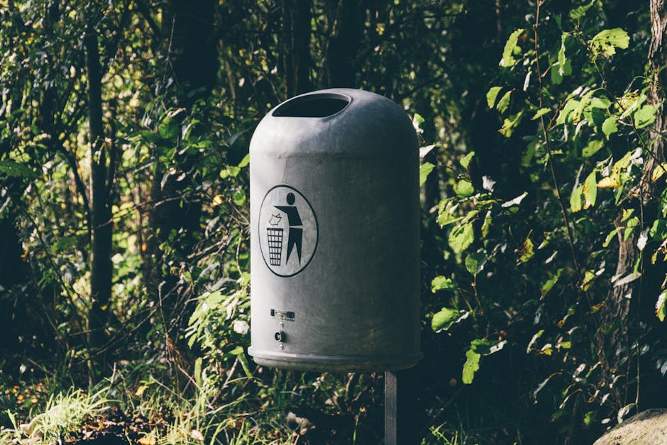 A grey, cylindrical outdoor rubbish bin made of metal, positioned on a metal post on a patch of grass. The bin features a rounded, slightly tapered top with an oval-shaped opening at its center for waste disposal. On its front, there is a black icon depicting a person throwing trash into a bin, along with a grid pattern representing the waste container. Surrounding the bin are dense green foliage, including leafy bushes and trees, with sunlight filtering through the leaves, creating dappled light on the ground and the bin. The scene suggests a natural setting, possibly a park or garden area, where outdoor waste collection objects are maintained for convenience. The presence of the bin indicates a focus on proper waste disposal in an outdoor environment, aligning with local rubbish removal or designated green waste management options often provided by waste collection services like Waste Collection Merton.
