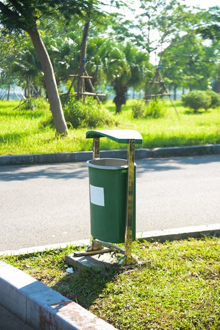 A green waste bin with a rectangular white label on the front, mounted on a metal frame with a curved, green plastic lid, situated on a grassy verge beside a paved roadway. The bin is positioned near the edge of the pavement, with the curb visible in the foreground. In the background, there are lush trees, including palm trees, and dense green foliage, suggesting a park or landscaped area. The scene is lit by natural daylight, with shadows cast by the trees and bin, indicating clear weather. The presence of the waste bin in this outdoor setting relates to waste management practices, with Waste Collection Merton offering local rubbish and waste disposal services, possibly including designated waste collection points for garden or green waste. The overall environment appears clean and well-maintained, emphasizing the importance of proper waste disposal in public outdoor spaces.