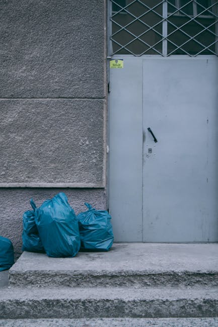 The image shows three blue plastic rubbish bags placed on a concrete step at the base of a grey exterior wall next to a metal door with a black handle and a keyhole. The wall has a textured finish, while the door appears smooth and painted in a light grey tone. Above the door, there is a section of metal grille covering a window or ventilation opening, with a darker background behind it. A small yellow and black warning sign is affixed near the top of the door frame. The environment suggests an urban or industrial setting, possibly a back entrance or service access point. The presence of the bags indicates waste awaiting collection, illustrating a scenario where private or independent rubbish disposal, such as through waste collection services like Waste Collection Merton, might be involved in managing refuse outside a building. The scene is lit evenly, with no strong shadows, providing a clear view of the materials and surroundings, fitting within the context of rubbish removal and waste management.