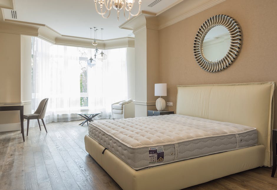 A spacious, well-lit bedroom featuring a large cream-colored upholstered bed with a new, white mattress on top. The bed is positioned against a beige wall decorated with a round, decorative mirror with a striped frame. To the right of the bed is a small black bedside table holding a white ceramic table lamp with a textured base and a white lampshade. On the left side of the room, there is a white armchair placed near large windows covered with sheer white curtains, allowing natural light to fill the space. The room has light wooden flooring and classic crown molding along the ceiling. In the foreground, a dark wooden side table and an upholstered chair are visible, completing the minimal and elegant interior. This scene reflects a typical bedroom that might be prepared for furniture disposal or replacement, in line with private waste handling services offered by Waste Collection Merton, emphasizing the importance of proper rubbish removal for large household items.