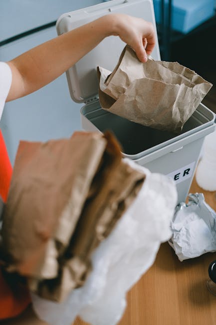 A person is seen disposing of waste by placing a crumpled brown paper bag into a grey and white rubbish bin with a partially open lid, situated on a wooden surface indoors. The person's arm is extended, holding the paper bag from the top, with their wrist visible and wearing a white sleeve. In the foreground, there is a stack of additional brown paper bags, some crumpled, which are awaiting disposal. The environment appears to be a home or office space, with a clean, well-lit setting, and the bin is positioned against a wall or cabinet. The scene illustrates typical aspects of household or commercial waste management, aligning with private or independent rubbish collection services. Waste Collection Merton's waste disposal solutions often include on-site clearance and alternative waste handling options, as demonstrated by the organized disposal of paper waste in this scenario, reinforcing the importance of proper rubbish management in compliance with local regulations.