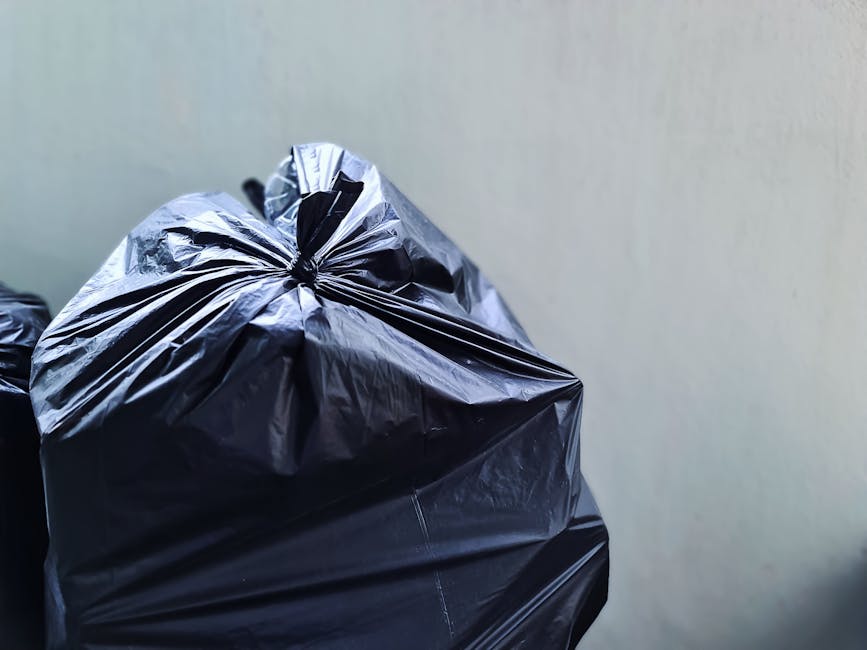 Close-up of a black plastic rubbish bag filled with waste, tied at the top with visible creases and reflective surface, resting on a surface in front of a plain, neutral-colored wall. The bag appears full and slightly bulging, with textured folds showing the plastic's sheen and crinkled material. In the background, there are additional black rubbish bags stacked or leaning against the wall, indicating an area designated for waste collection or temporary storage, possibly on a driveway or outdoor space. The environment is lit with natural or ambient lighting, highlighting the shiny surface of the plastic and the surrounding context of waste handling, which relates to private or independent rubbish disposal services like those offered by Waste Collection Merton in accordance with local rubbish management regulations.