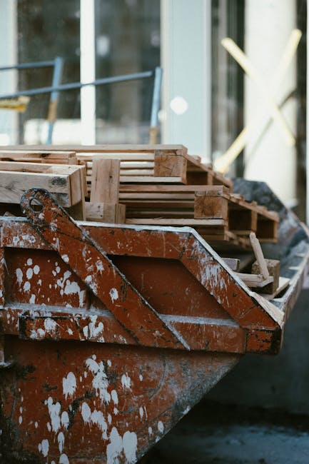 A close-up view of a rusty orange metal boat filled with freshly assembled wooden pallets, some with visible nailing and rough edges, resting against a dark body of water. The pallets are light-colored, untreated wood with smooth, flat surfaces and slatted designs, stacked neatly but unevenly within the boat. The boat appears to be docked or positioned on the water's edge, with a blurred background showing a modern glass building with reflective surfaces and architectural elements like scaffolding and a large X-shaped structure. The environment indicates an outdoor setting, possibly a construction site or renovation project involving the removal of wooden debris or pallet waste. The lighting is natural, with soft diffuse daylight illuminating the scene, emphasizing the textures of the weathered metal and fresh wood, aligning with themes of on-site clearance or alternative waste handling by waste collection services such as Waste Collection Merton.