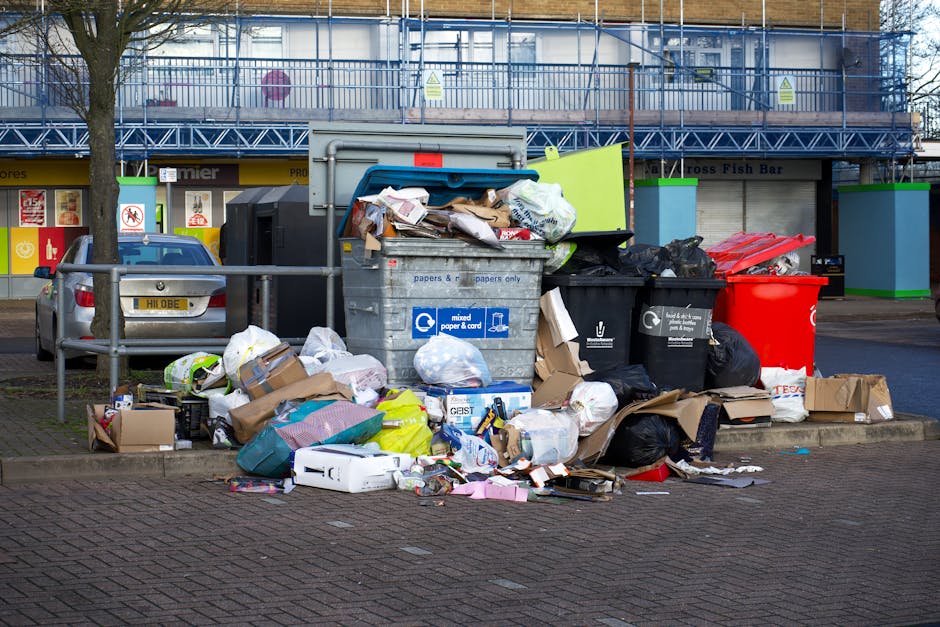A collection of overflowing rubbish bins and scattered trash on a paved urban sidewalk, featuring a large grey mixed paper and card waste container with its lid open and bulging with various crumpled paper, cardboard boxes, and plastic packaging. Surrounding the container are numerous black, white, and red garbage bags, some torn open, with loose packaging, cardboard boxes, and discarded items strewn across the ground. Behind the clutter, there is a parked silver car with a yellow license plate, positioned adjacent to a metal railing. In the background, a row of small commercial units with brightly coloured signage and a building under construction or renovation with a blue scaffold are visible, indicating an area where private or independent waste collection such as that offered by Waste Collection Merton may be necessary for on-site waste management or additional rubbish removal services beyond standard council collection. The image is set in daylight, with natural lighting highlighting the chaotic scene of refuse in an urban environment.