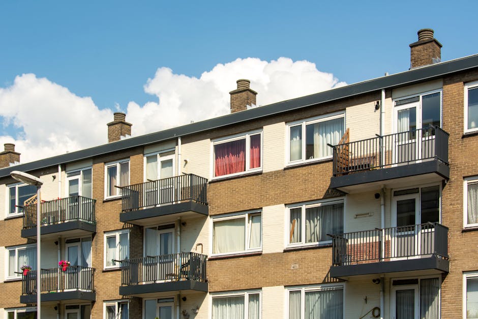 A three-story residential apartment building constructed with brown and cream brickwork, featuring multiple balconies with black metal railings on the front facade. Each balcony has different items, such as chairs and flower pots, placed on them. The windows are largely white-framed, with some curtains visible, including red and cream colors. The building's flat roof is topped with several brick chimneys, and a streetlight is partially visible to the left. The image is taken during the daytime with a bright blue sky and scattered white clouds in the background. This scene depicts a typical urban residential complex, where the exterior conditions suggest routine maintenance, and the building is suitable for private clearance services or rubbish removal of outdoor clutter or debris in surrounding common areas, aligning with waste collection practices in private housing estates.