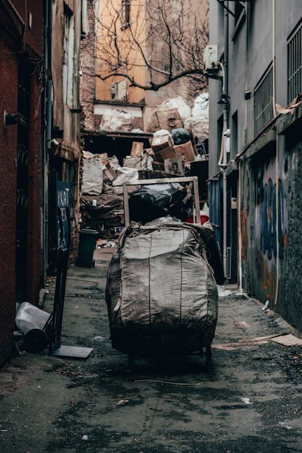 A collection of overflowing rubbish bins and scattered trash on a paved urban sidewalk, featuring a large grey mixed paper and card waste container with its lid open and bulging with various crumpled paper, cardboard boxes, and plastic packaging. Surrounding the container are numerous black, white, and red garbage bags, some torn open, with loose packaging, cardboard boxes, and discarded items strewn across the ground. Behind the clutter, there is a parked silver car with a yellow license plate, positioned adjacent to a metal railing. In the background, a row of small commercial units with brightly coloured signage and a building under construction or renovation with a blue scaffold are visible, indicating an area where private or independent waste collection such as that offered by Waste Collection Merton may be necessary for on-site waste management or additional rubbish removal services beyond standard council collection. The image is set in daylight, with natural lighting highlighting the chaotic scene of refuse in an urban environment.