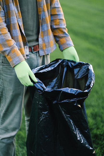 A person dressed in casual clothing, including a plaid shirt with blue, yellow, and grey tones, and grey trousers, is seen standing outdoors on a grassy area. The individual is wearing light green gloves and holding open a large black rubbish bag made of coated plastic, which is partially expanded and positioned upright. Behind them, the background features green grass and blurred natural vegetation, suggesting a garden or park setting. The scene appears to depict waste collection or disposal activity, consistent with professional rubbish removal practices. The person’s posture indicates they are ready to insert waste materials into the bag, aligning with services such as on-site clearance or private waste handling offered by waste collection specialists like Waste Collection Merton, operating in the vicinity of Merton. The natural outdoor environment and the practical use of gloves and a large rubbish bag reflect a typical scene associated with rubbish or waste management tasks meant for efficient disposal or collection outside of conventional municipal services.
