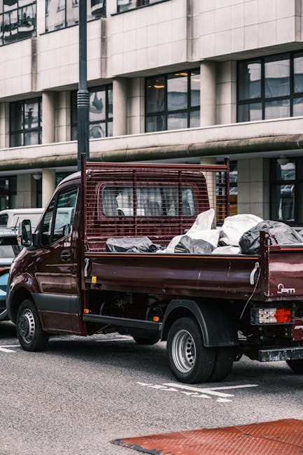 A compact, dark red flatbed truck parked on a city street in front of a modern office building with large rectangular windows. The truck's cargo area is loaded with various types of rubbish, including several white and grey bags filled with waste, some of which are partially open revealing indistinct contents. The truck features a metal mesh protective panel behind the cab, providing safety and support for securing the load, and the tailgate is lowered to accommodate the debris. The vehicle is situated on the roadway beside a designated parking area, with some other vehicles visible in the background. The environment appears urban, with ambient daylight and a slightly overcast sky, highlighting the practicality of private waste removal, such as that offered by Waste Collection Merton, for managing rubbish in commercial or residential settings without relying solely on local authority services.