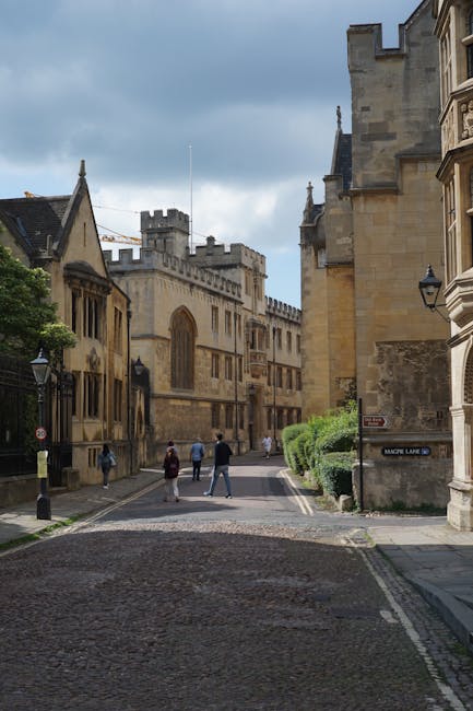 A narrow cobbled street in an historic city area, lined with both Gothic and classical stone buildings with detailed facades, large windows, and decorative stonework. On the right, a sandstone building features ornate architectural details and a black street sign reading 'Magdalen Lane.' On the left, a larger structure with crenellated towers and battlements suggests a medieval or university building, with tall, arched windows and ornate stonework. Several pedestrians walk along the street, some in pairs and others alone, dressed in casual clothing suitable for cool weather. Small, neatly trimmed green bushes grow at the base of the buildings on the right, adding a touch of greenery to the stone environment. The sky above is partly cloudy with patches of blue visible, casting soft daylight that highlights the textures and warm tones of the historic stonework. This scene reflects a typical setting for historic city centres, where independent or private waste collection might be relevant for maintaining building facades and streetscapes, with Waste Collection Merton providing such services in similar urban environments.