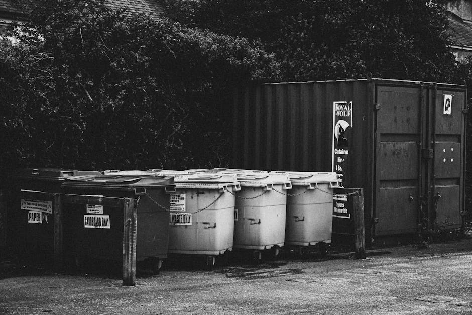 A row of five large, white plastic wheeled waste bins with closed lids is positioned on a paved surface outdoors. Each bin is marked with black labels indicating specific waste types, such as 'ELECTRONIC WASTE ONLY' and 'GENERAL WASTE,' and they are arranged side by side, aligned parallel to a dark, corrugated metal storage container in the background. The containers are situated near a weathered wooden post with additional signage for waste disposal, and behind them are dense trees and foliage, suggesting a residential or private property boundary. The environment appears calm and well-maintained, with natural light illuminating the scene evenly, emphasizing the textures of the plastic bins, the metal container, and the surrounding shrubbery. This setup reflects a typical arrangement for an independent waste collection or on-site rubbish disposal area managed by Waste Collection Merton, suitable for handling various waste streams in accordance with local waste management practices.